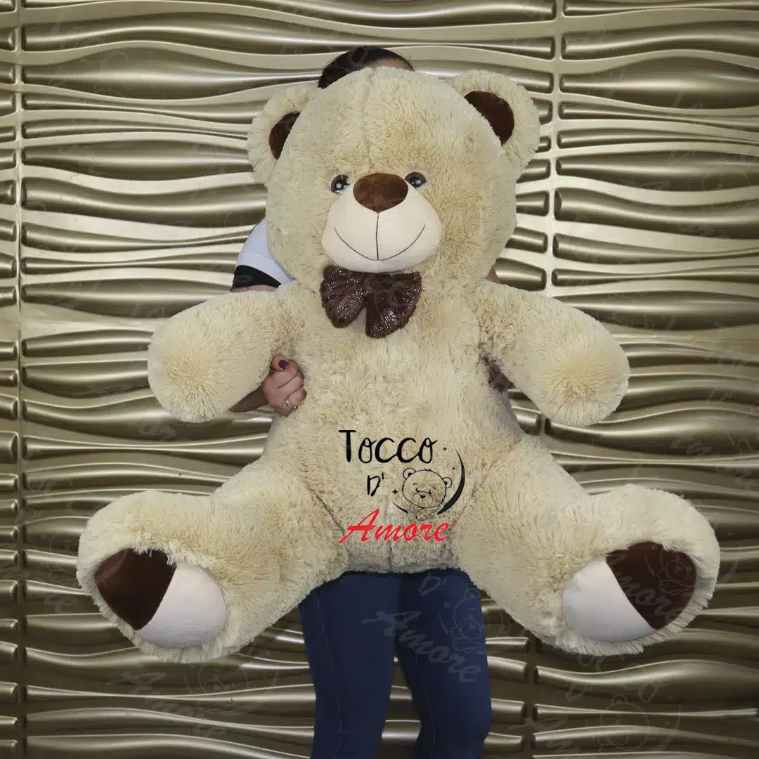 Oso de peluche grande beige de 1,10 m con moño café, sostenido frente a una pared decorativa
